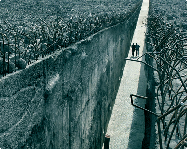 Zdjęcie. Dwie osoby idą chodnikiem. Po bokach wysokie ściany zakończone drutami 