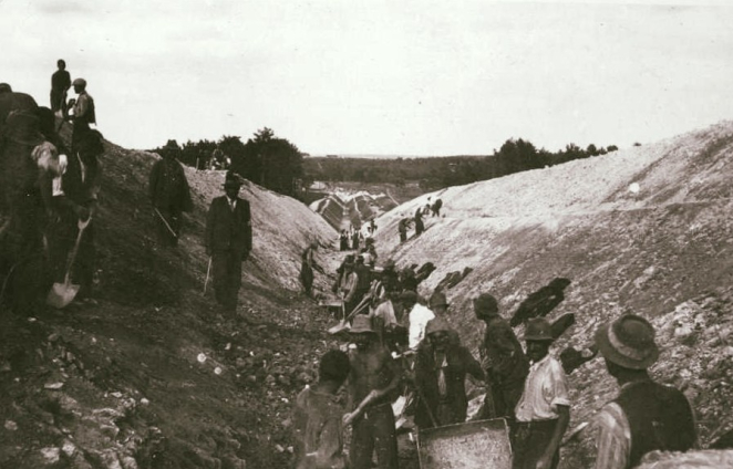 Grupa kilkudziesięciu osób z łopatami pośrodku podłużnego rowu.
