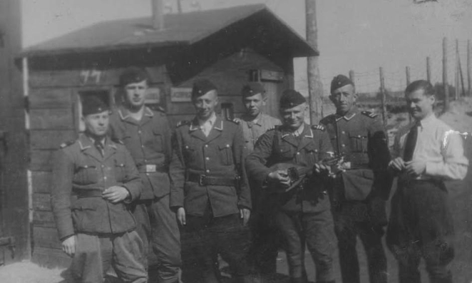 Siedmiu mężczyzn w mundurach, jeden z mężczyzn z mandoliną, w tle drewniany mały budynek.