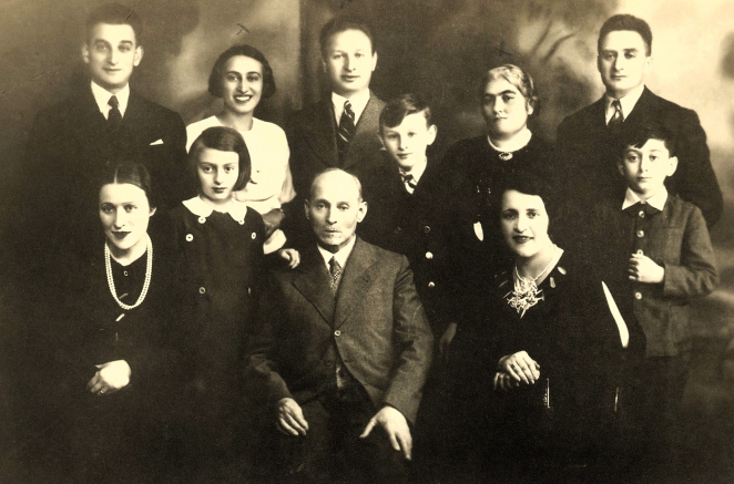 Zdjęcie w sepii, portret jedenastoosobowej rodziny; w pierwszym rzędzie na środku starszy mężczyzna w garniturze, obok dwie ciemnowłose kobiety ubrane na czarno z naszyjnikami z pereł, pomiędzy nimi dzieci (dziewczynka i dwóch chłopców), ostatni rząd, trzech mężczyzn i dwie kobiety - jedna starsza ubrana na czarno, młodsza uśmiechnięta ubrana na biało. 