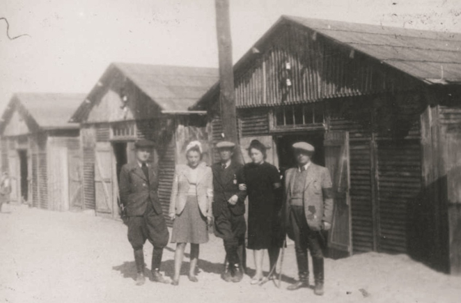 Grupa pięciu osób (dwie kobiety, trzech mężczyzn) ubranych w cywilne ubrania na lewej ręce widoczna jasna opaska, na drugim planie rzędy drewnianych baraków. 