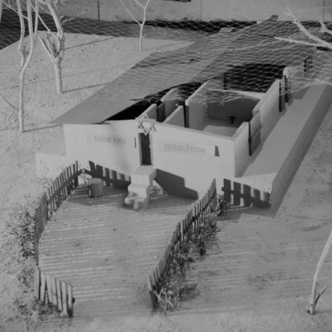 Prostokątny model budynku, drewniany płot przez wejściem, napis: Bade und Inhalation oraz znak: Gwiazda Dawida, dach częściowo odsłonięty ukazujący prostokątne pomieszczenia, dookoła drzewa bez liści. 