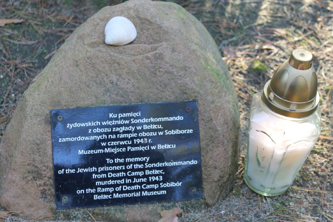 Kamień z przymocowaną do niego czarną tabliczką z napisem: Ku pamięci żydowskich więźniów Sonderkommando z obozu zagłady w Bełżcu zamordowanych na rampie obozu w Sobiborze w czerwcu 1943 r. Muzeum-Miejsce Pamięci w Bełżcu. Pod spodem angielskie tłumaczenie napisu. Po lewo biały znicz, na kamieniu ułożony biały mały kamień. 