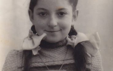 Czarno-białe zdjęcie portretowe dziewczynki, Bracha (Broni) Beer-Rauffmann, córki Salomei, z warkoczami przewiązanymi jasnymi kokardami, patrzącej w obiektyw na jasnym tle.