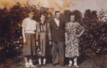 Zniszczona, powojenna fotografia przedstawiająca rodzinę Brogowskich z córkami stojących razem w ogrodzie na tle krzewów.