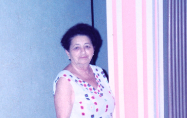 Kolorowe zdjęcie Salomei Beer stojącej przy ścianie w pasy, w jasnej sukience w drobny wzór, otoczonej bukietami kwiatów.