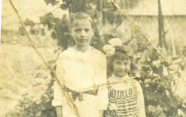 Czarno-biała fotografia z 1943 roku przedstawiająca Marię Brogowską i Irenę Sznycer stojące razem w ogrodzie, w jasnych dziecięcych strojach.