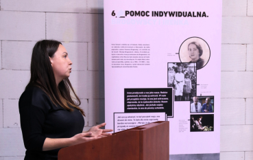 Kobieta przemawia przy mównicy podczas otwarcia wystawy w Muzeum i Miejscu Pamięci w Bełżcu, na tle planszy ekspozycyjnej.