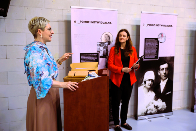 Kobieta przemawia przy mównicy podczas wydarzenia, a druga stoi obok na tle plansz ekspozycyjnych.