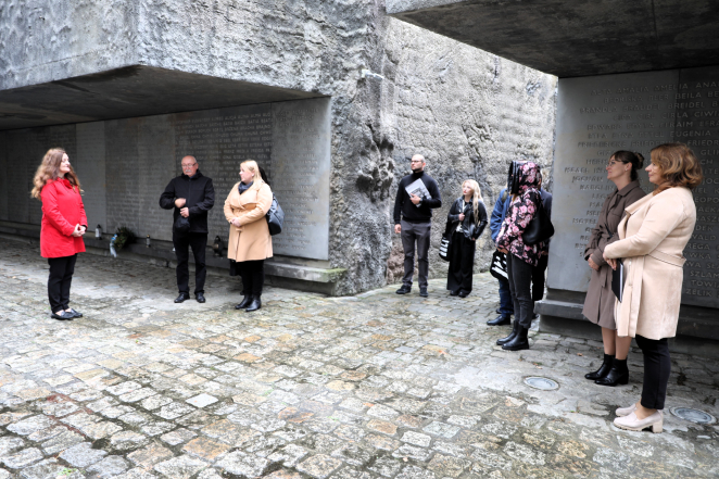 Grupa osób uczestniczy w spotkaniu upamiętniającym przy ścianach z nazwiskami ofiar w przestrzeni memorialnej.