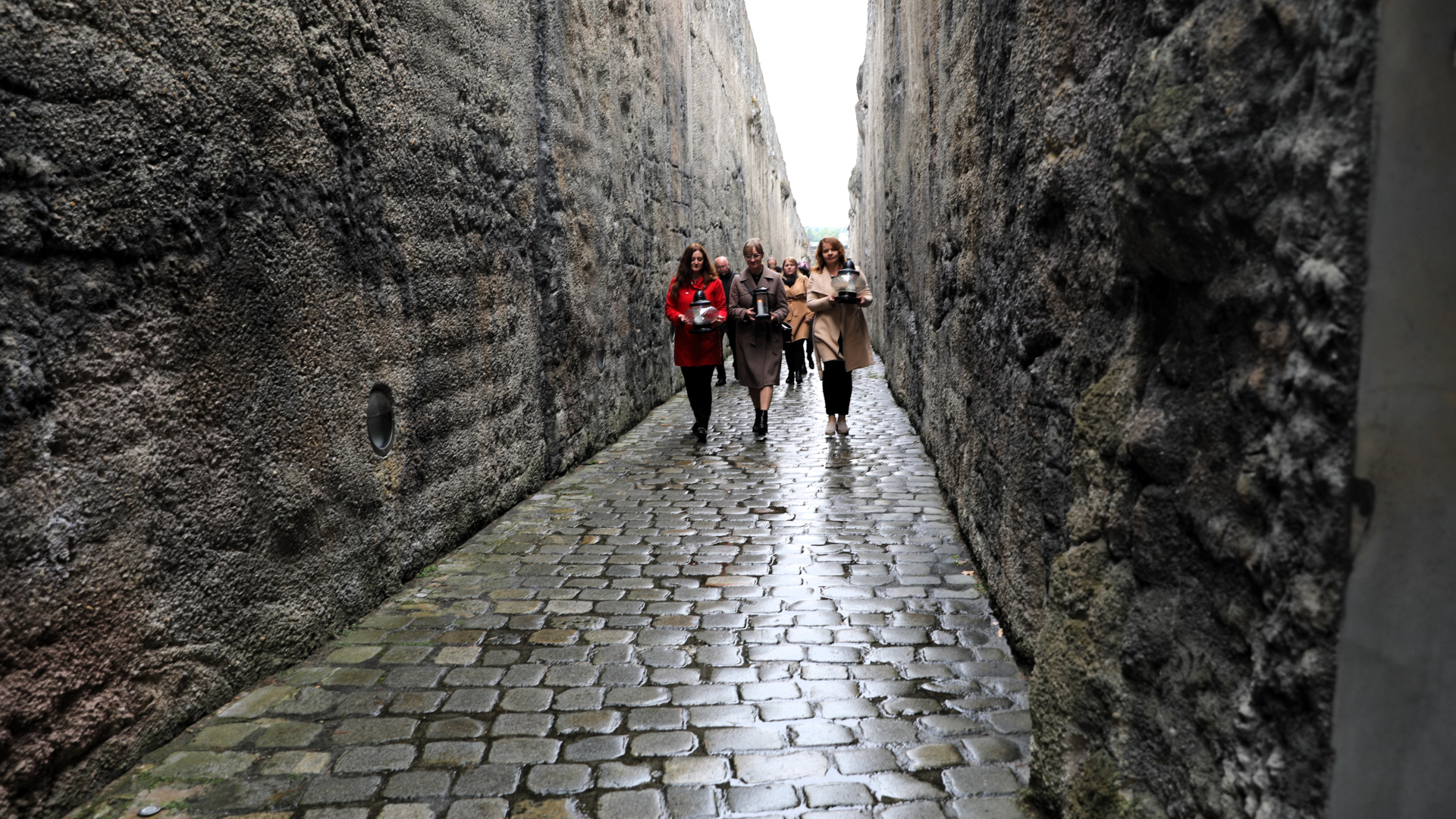 Grupa osób idzie z kwiatami przez wąską szczelinę pomnika, otaczają je ogromne betonowe ściany.