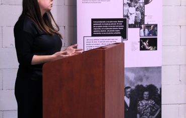 Kobieta przemawia przy mównicy, na tle planszy ekspozycyjnej.