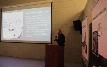 Prelegent przemawia przy mównicy podczas seminarium, na tle slajdu prezentacji i plansz wystawy.
