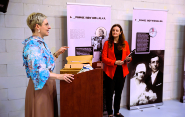 Kobieta przemawia przy mównicy podczas seminarium, a druga stoi obok na tle plansz ekspozycyjnych.