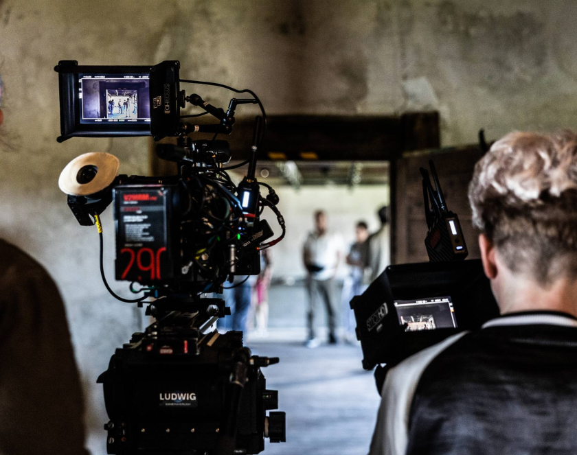 Photo. Two people are standing inside a building with their backs turned. Cameras are positioned in front of them. In the background, there are indistinct figures.