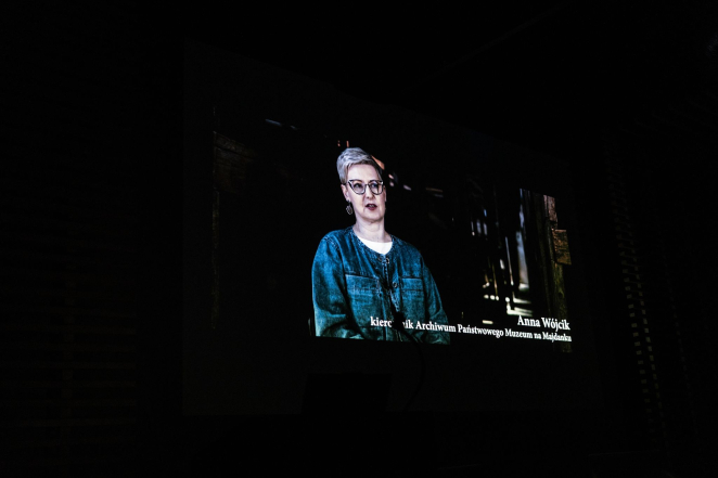 Dark photograph. In the center, there is a still from a film showing a woman wearing glasses. Caption: Anna Wójcik, Head of the Archives of the State Museum at Majdanek.