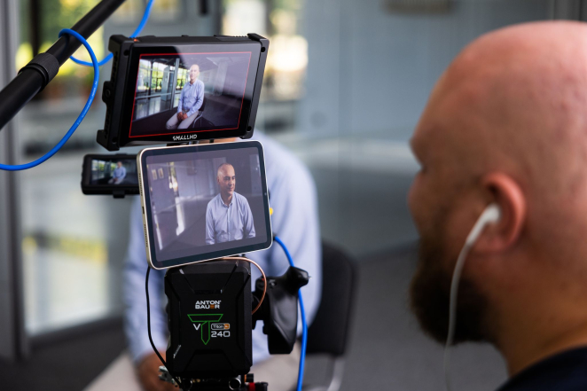 In the photo, there are the displays of a camera and a phone showing a man dressed in light-colored clothing. On the right, the head of a bald man with an earpiece in his ear.