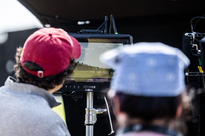 Photo. Two people are standing with their backs turned. In front of them, there is a camera screen showing indistinct figures next to prisoner barracks.