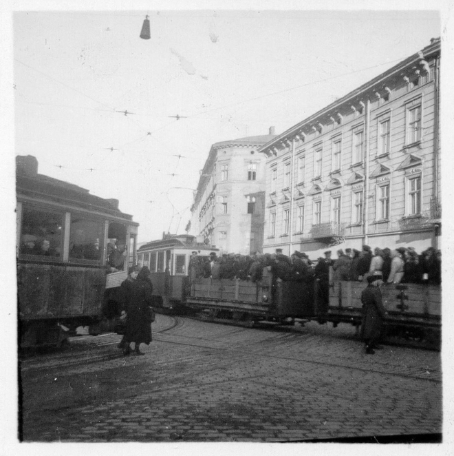 Czarno-białe zdjęcie historyczne przedstawiające stare wagony tramwajowe na brukowanej ulicy we Lwowie, z których dwa wypełnione są tłumem ludzi na tle miejskich kamienic.