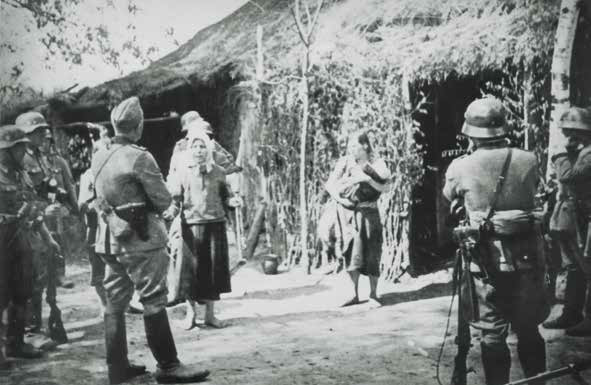 Czarno-biała fotografia. Na podwórku przed wiejską chatą krytą strzechą stoi grupa uzbrojonych niemieckich żołnierzy w hełmach i mundurach. Pomiędzy nimi znajdują się cywile, w tym bosa kobieta w chuście oraz inna kobieta trzymająca na rękach małe dziecko