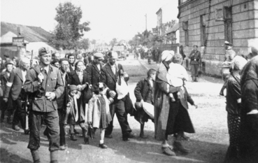 Fotografia przedstawia kolumnę cywilów idących ulicą obok piętrowego budynku. Ludzie niosą bagaże, niektórzy idą z dziećmi. Po lewej stronie kolumny kroczy uzbrojony mężczyzna w mundurze.