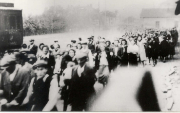 Lekko nieostra fotografia przedstawiająca duży tłum ludzi idących wzdłuż drogi, obok stojącego po lewej stronie ciemnego wagonu kolejowego.