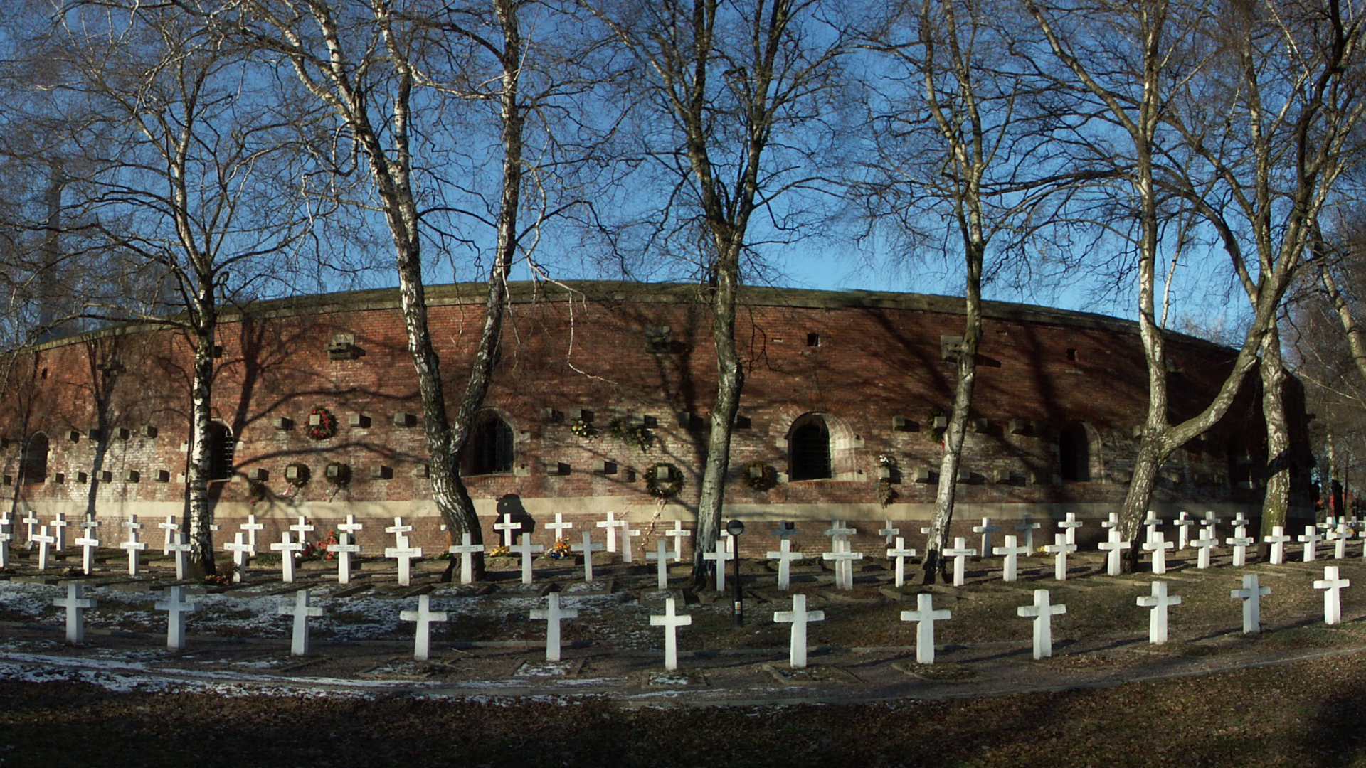 Panorama przedstawia Rotundę Zamojską – ceglaną budowlę obronną na planie koła, która podczas II wojny światowej służyła jako miejsce kaźni. Na pierwszym planie, wokół monumentalnych murów z ciemnej cegły, rozciąga się cmentarz z rzędami prostych, białych krzyży, stojących na trawiastym terenie. Między grobami rosną wysokie, bezlistne brzozy.