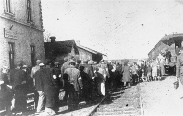 Fotografia przedstawia dużą grupę osób na stacji kolejowej, wszyscy odwróceni są tyłem. Niektórzy mają na rękawach białe opaski. Po prawej stronie jest mężczyzna w mundurze oraz wagon kolejowy, do którego wsiadają ludzie, natomiast po lewej stronie znajduje się fragment budynku stacji.