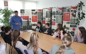 Zajęcia edukacyjne „Kronikarze czasów nieludzkich” w sali bibliotecznej. Grupa młodzieży siedzi przy długim, jasnym stole, słuchając mężczyzny w niebieskiej koszuli. W tle, za uczestnikami, ustawionych jest kilka wysokich plansz wystawowych