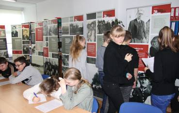 Młodzież podczas pracy warsztatowej w bibliotece. Na pierwszym planie dwie dziewczyny siedzą przy stole i wypełniają arkusze zadań. Obok nich oraz w tle pozostali uczestnicy stoją przy planszach wystawowych, uważnie czytając teksty.