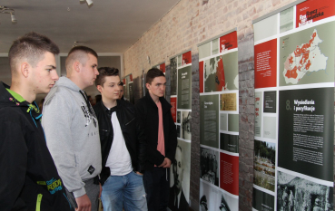 Czterech młodych mężczyzn stoi wewnątrz pomieszczenia o ścianach z surowej, czerwonej cegły. Wszyscy przyglądają się wielkoformatowym planszom edukacyjnym, które stanowią element wystawy historycznej.