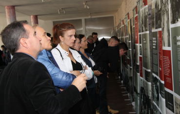 Grupa dorosłych osób ogląda wystawę historyczną „Res humana – rzecz nieludzka”. Postacie na pierwszym planie przyglądają się rzędowi pionowych, biało-czerwono-szarych planszy. Ekspozycja, ustawiona wzdłuż ściany z surowej cegły, zawiera liczne archiwalne fotografie, mapy oraz bloki tekstu dotyczące okupacji Zamojszczyzny.