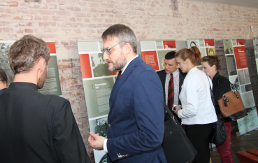 Zdjęcia dokumentują wernisaż wystawy o okupacji Zamojszczyzny. Widzowie w różnym wieku oglądają biało-czerwone plansze z mapami i fotografiami archiwalnymi, rozwieszone na tle surowej, ceglanej ściany.