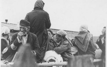 Fotografia przedstawia grupę osób na wozie. Na pierwszym planie widać starszego mężczyznę w ciemnej czapce, a wokół niego kobiety w chustach na głowach, niektóre odwrócone tyłem.