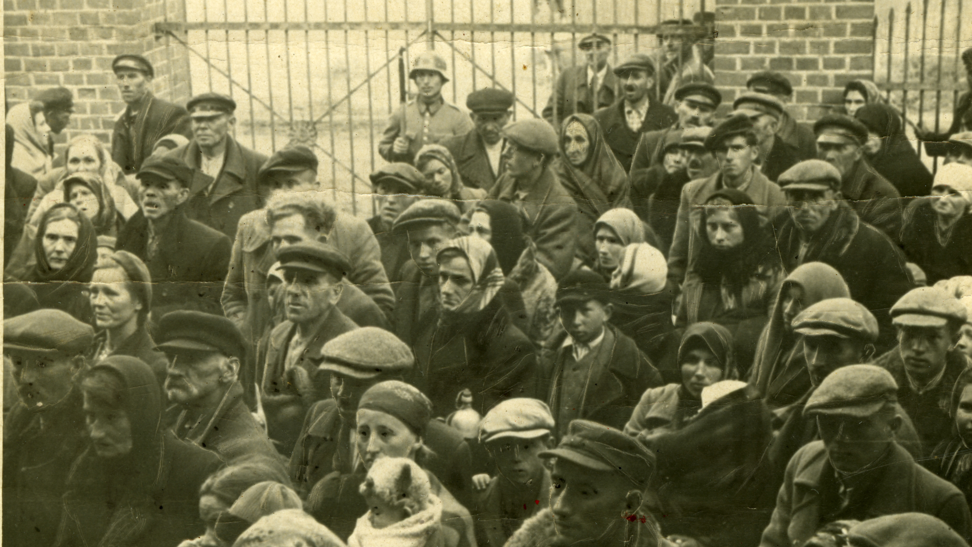 Archiwalne, czarno-białe zdjęcie o sepiawym odcieniu, przedstawiające tłum cywilów podczas akcji wysiedleńczej. Na pierwszym planie widać stłoczone osoby w płaszczach, kaszkietach i chustach na głowach, których twarze malują niepokój i zmęczenie. W tle, przed metalowym ogrodzeniem, dostrzec można sylwetkę uzbrojonego niemieckiego żołnierza, pilnującego zgromadzonych.