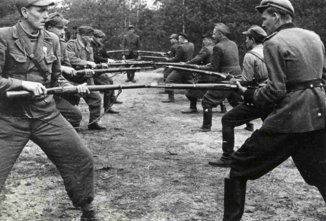 Czarno-białe zdjęcie przedstawiające szkolenie partyzanckie w powiecie biłgorajskim. Na leśnej polanie dwie grupy mężczyzn w mundurach stoją naprzeciw siebie w rozkroku, ćwicząc walkę wręcz. Każdy z nich trzyma oburącz karabin z nasadzonym bagnetem, celując w stronę przeciwnika.