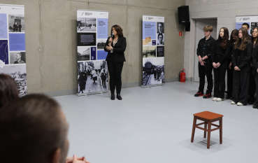 Współczesna fotografia przedstawiająca kobietę przemawiającą do mikrofonu w sali wystawowej. Wokół niej stoją plansze wystawy czasowej ze zdjęciami historycznymi i tekstami, a z boku przygląda się grupa młodych ludzi.