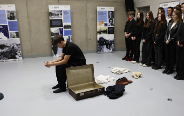 Współczesne zdjęcie. Na środku jasnej sali siedzi tyłem do widzów młody mężczyzna w czarnym ubraniu, pochylony obok otwartej, pustej walizki. Obok niego na podłodze leżą rozrzucone przedmioty: buty, części garderoby oraz czapka. W tle, pod ścianą, stoi grupa młodzieży obserwujących scenę, a obok nich widoczne są edukacyjne plansze z historycznymi zdjęciami i tekstami.