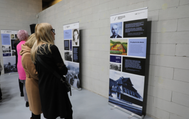 Współczesna fotografia przedstawiająca trzy kobiety oglądające plansze wystawy o deportacjach do obozu zagłady w Bełżcu. Kobiety stoją tyłem do obiektywu w jasnej sali z betonowymi ścianami.
