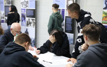 Współczesna fotografia przedstawiająca grupę młodzieży i starszego mężczyznę pracujących wspólnie przy stoliku w sali edukacyjnej. W tle są plansze wystawowe z historycznymi zdjęciami i tekstami, przy których stoją inne osoby.