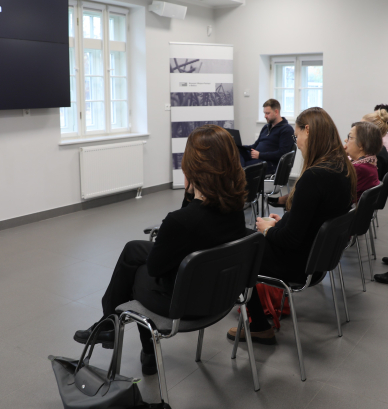 Na zdjęciu widać prelekcję w jasnej, nowoczesnej sali. Po lewej stronie stoi prowadząca przy dużym ekranie z napisem „obóz pracy – geneza”, a naprzeciwko niej siedzi grupa zasłuchanych osób.