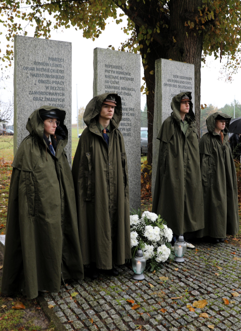 Czworo harcerzy w zielonych pelerynach przeciwdeszczowych pełni wartę przed trzema kamiennymi tablicami upamiętniającymi ofiary Romów i Sinti. U stóp pomnika, w jesiennej scenerii, złożono białe kwiaty i zapalono znicze.