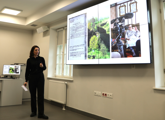 Prelegentka prowadzi prezentację w sali konferencyjnej; na dużym ekranie wyświetlone są slajdy z dokumentem, krajobrazem i nagraniem wywiadu