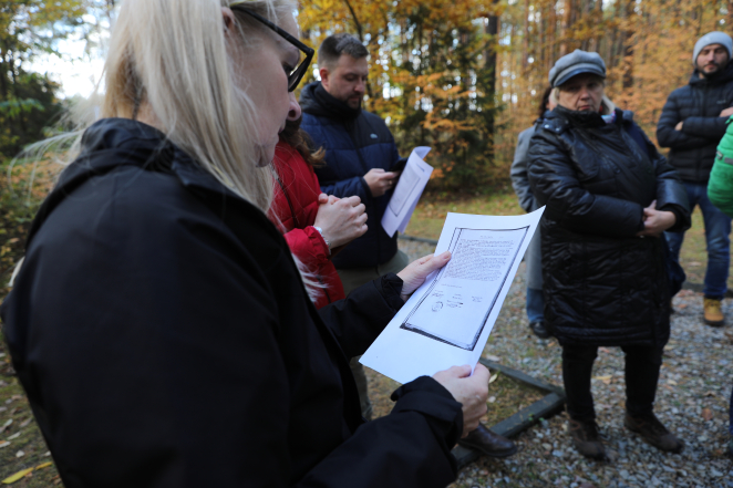 Kobieta w czarnej kurtce uważnie czyta kopię archiwalnego dokumentu podczas jesiennego spotkania w lesie, w którym uczestniczy grupa osób skupionych na historycznych przekazach.