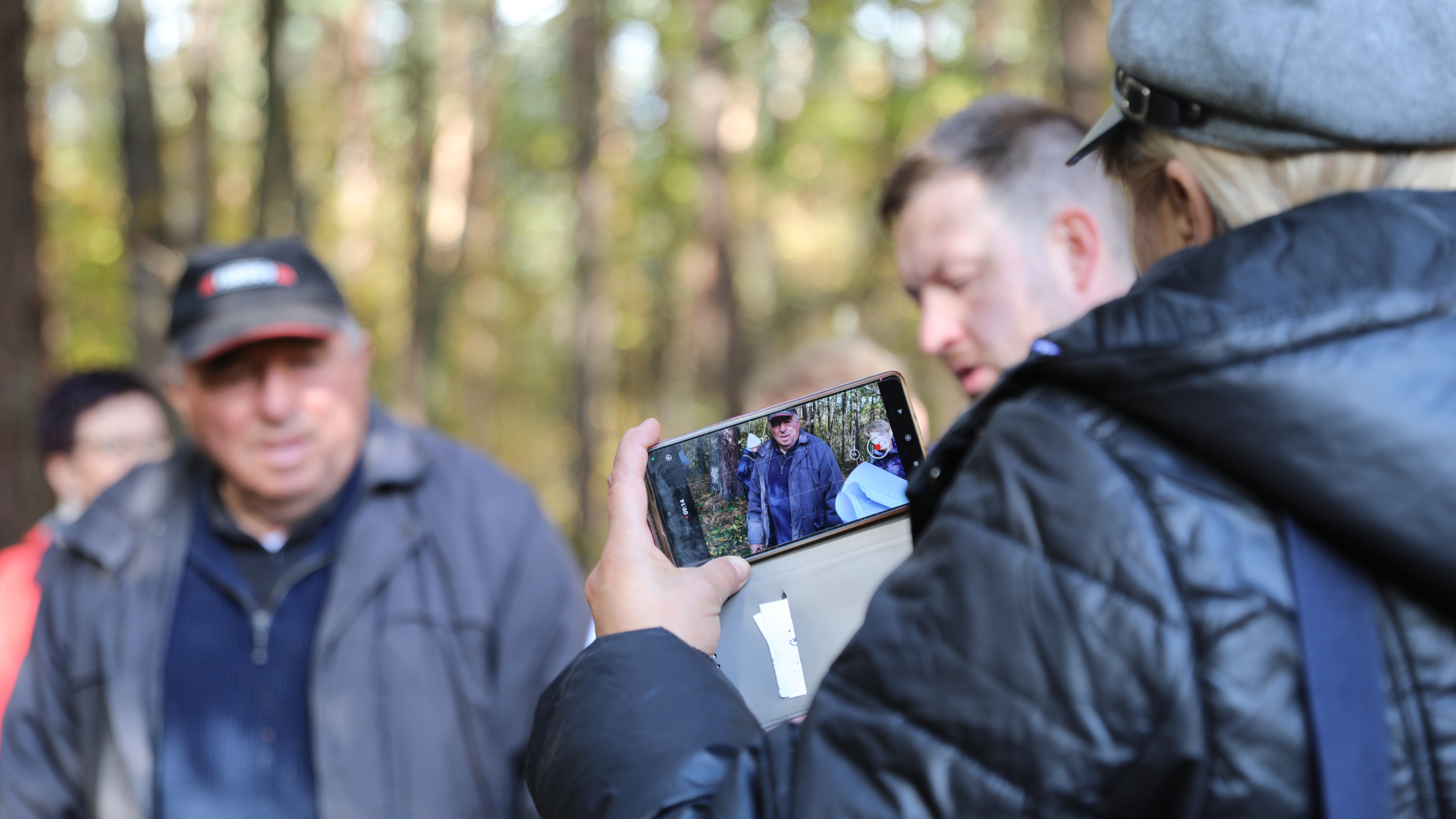 Ludzie zgromadzeni w lesie słuchają świadka historii. Jedna z uczestniczek nagrywa przemawiającego mężczyznę swoim telefonem.