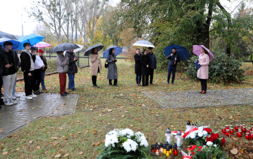 Uczestnicy uroczystości upamiętniającej zgromadzili się pod parasolami w deszczowy, jesienny dzień . Grupa osób w różnym wieku słucha kobiety w różowym płaszczu, która przemawia, trzymając folder z dokumentami. Na pierwszym planie, na trawie zasłanej liśćmi, widoczne są złożone wiązanki kwiatów z biało-czerwonymi wstążkami oraz liczne zapalone znicze.