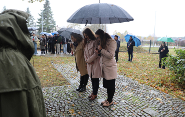 Trzy kobiety w płaszczach, chroniąc się pod wspólnym czarnym parasolem, stoją z pochylonymi głowami na brukowanej ścieżce, oddając hołd ofiarom.