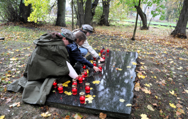 Trzy młode harcerki klęczą przy płycie pamięci w lesie i składają zapalone znicze.