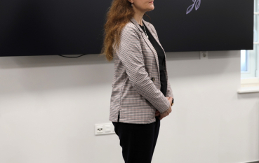 Kobieta w kraciastej marynarce stoi obok dużego ekranu z napisem: „Historia i pamięć: 85. rocznica zagłady Romów i Sinti w obozie pracy w Bełżcu”. Spotkanie odbywa się w nowoczesnej sali edukacyjnej muzeum.