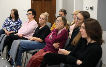 Grupa kobiet siedzi w rzędach, z uwagą i powagą przysłuchując się prowadzonej prelekcji historycznej.